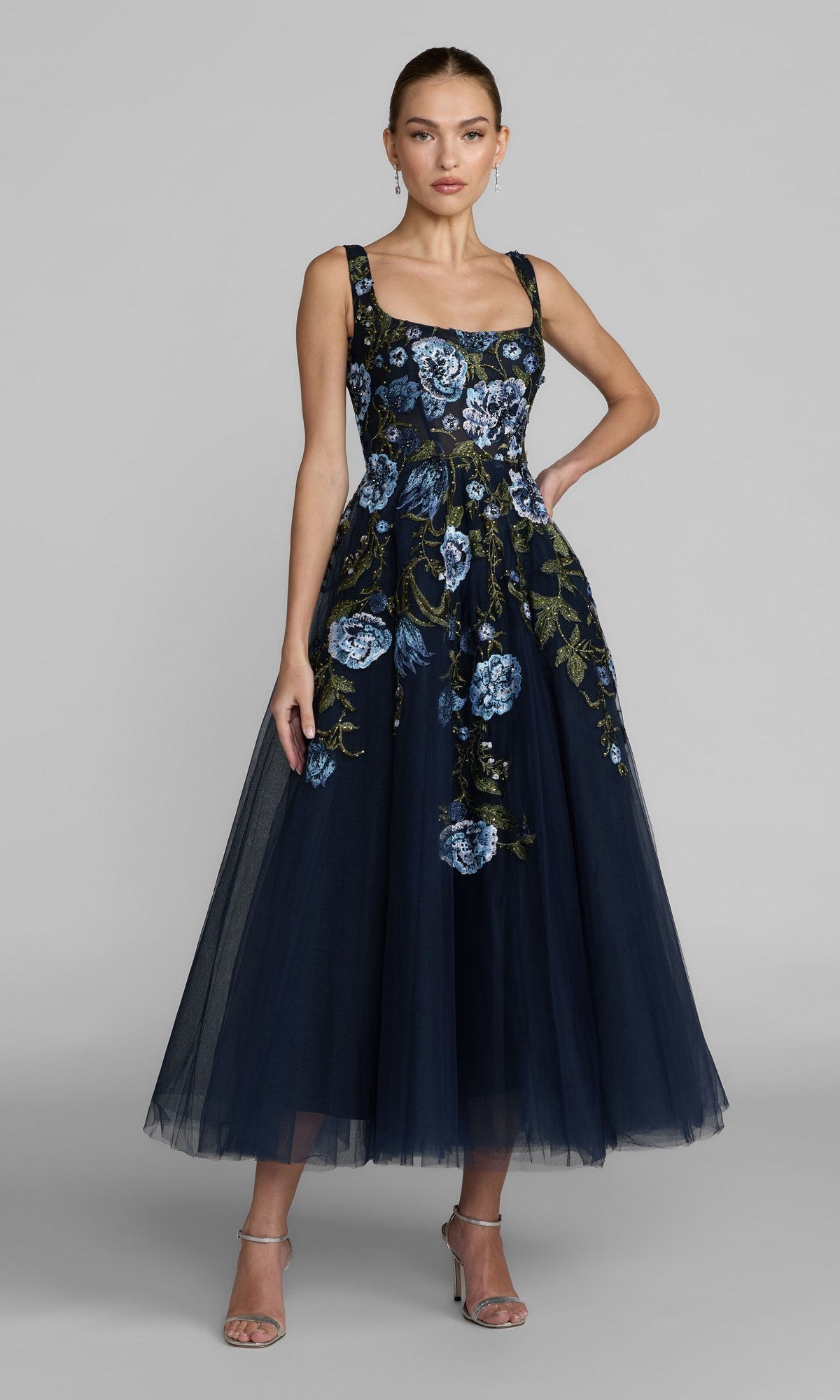 A woman stands against a plain gray background, her hair pulled back, wearing the Mac Duggal 21295 Tea-Length Party Dress in Blue Multi with sleeveless styling and beautiful blue and green floral embroidery, paired with silver strappy heels.